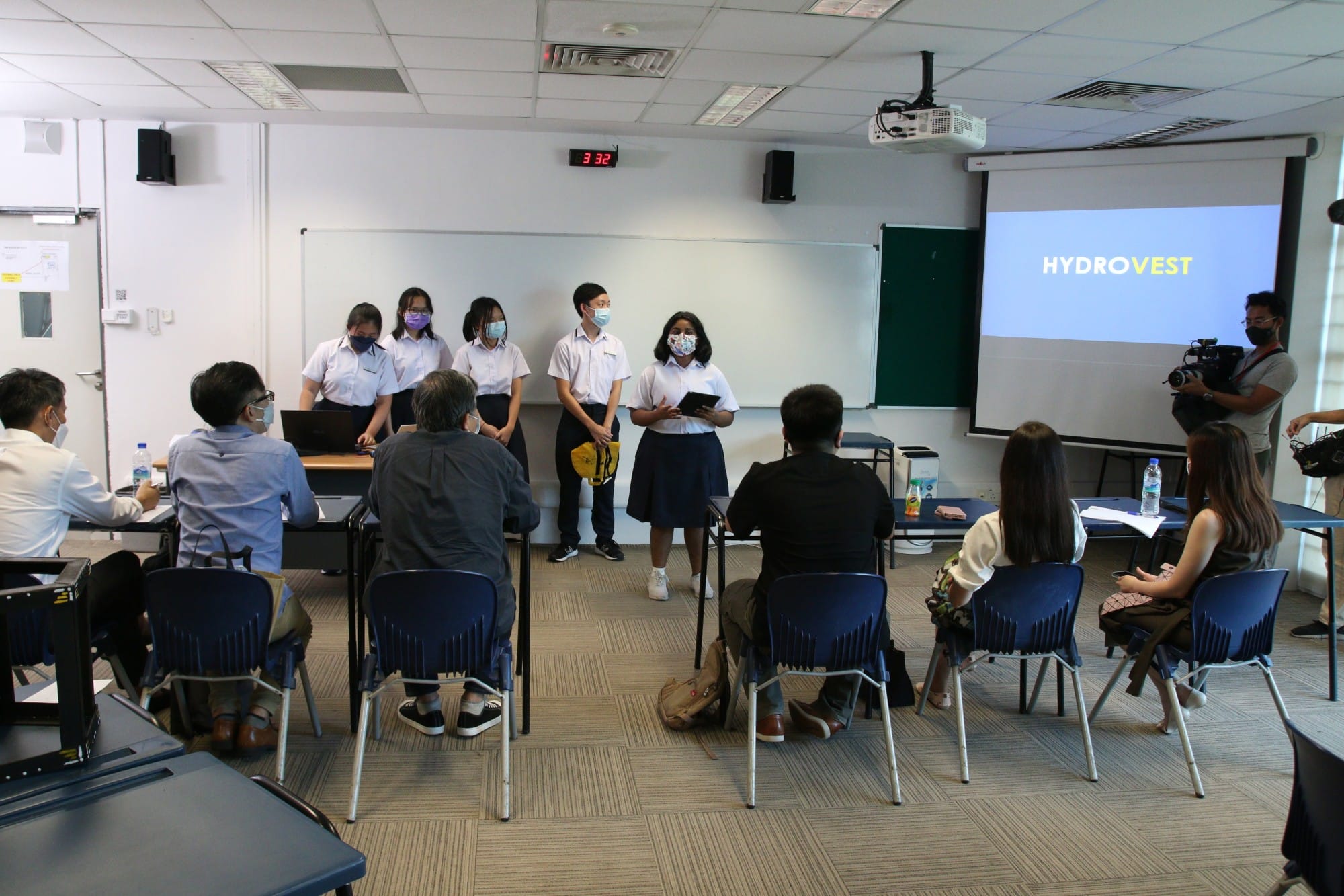 Four masked students in uniform present "HYDROVEST" on a projected screen to seated judges in a classroom.